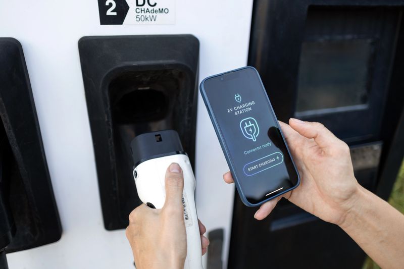 Hands holding an ev charger and a smartphone at a charging station, controlling the charging process with a mobile app