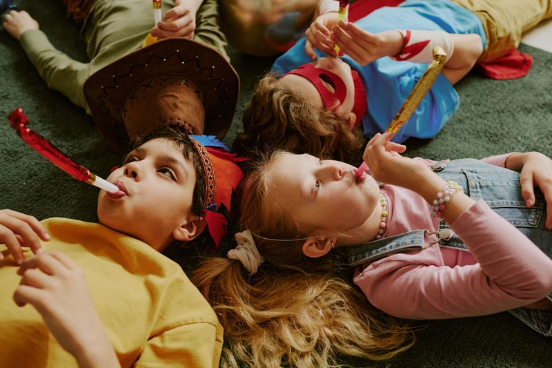 Group of multiethnic children lying on floor blowing party horns, wearing costumes and accessories, celebrating together during festive event, showing playful expressions and relaxed poses