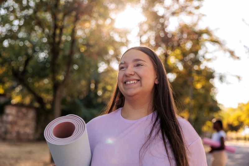 Smiling young woman carrying an exercise mat while walking outdoors. She looks happy and full of positive energy, representing a healthy lifestyle, fitness, and well-being.