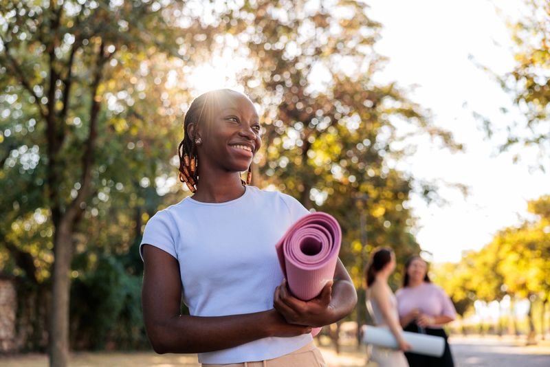 Smiling young black woman walking outdoors with an exercise mat in her hands, full of positive energy. Behind her, two friends are chatting and also carrying exercise mats, symbolizing friendship, fitness, and a healthy lifestyle.