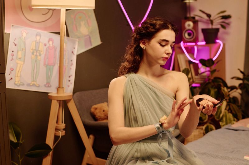 Caucasian young adult woman sitting in bedroom applying floral perfume, surrounded by plants and artwork, wearing elegant dress, looking down at hands