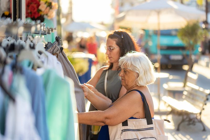 Two women looking through colorful clothes on a rack at a seaside market.