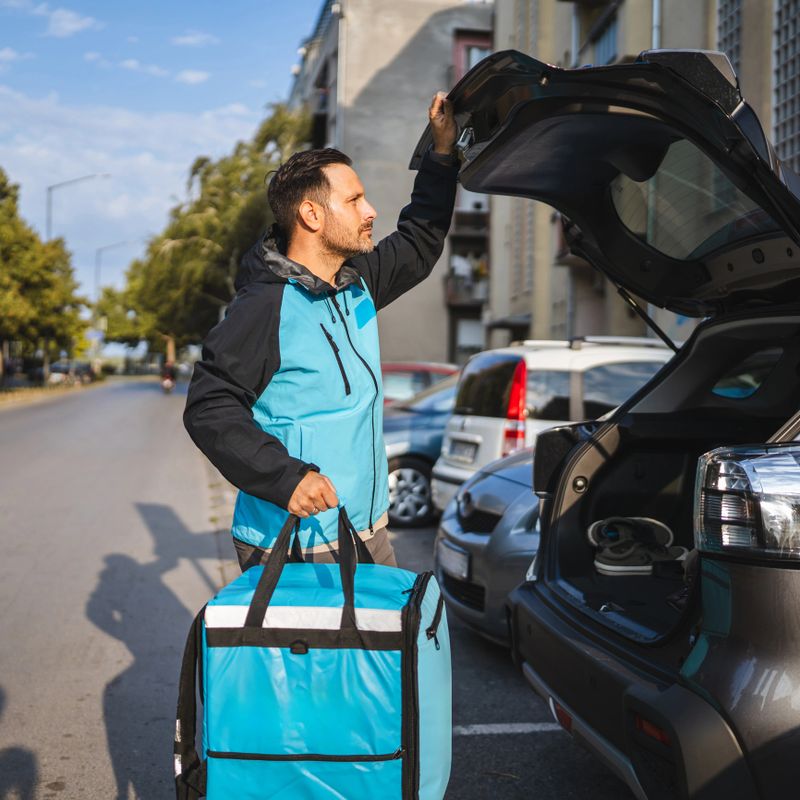 Delivery driver in uniform jacket opening car trunk and holding a thermal bag while getting ready for food delivery on a bustling city street, showcasing urban logistics in action