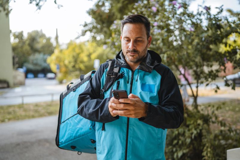 Delivery man wearing a thermal backpack, checking the next delivery address on a smartphone app while walking through the bustling city streets, navigating efficiently to fulfill online orders