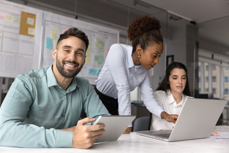 Diverse team collaborate on task gather in conference room, focus on smiling Hispanic man sitting at desk with digital tablet, look at camera exuding leadership and satisfaction, using innovative tech