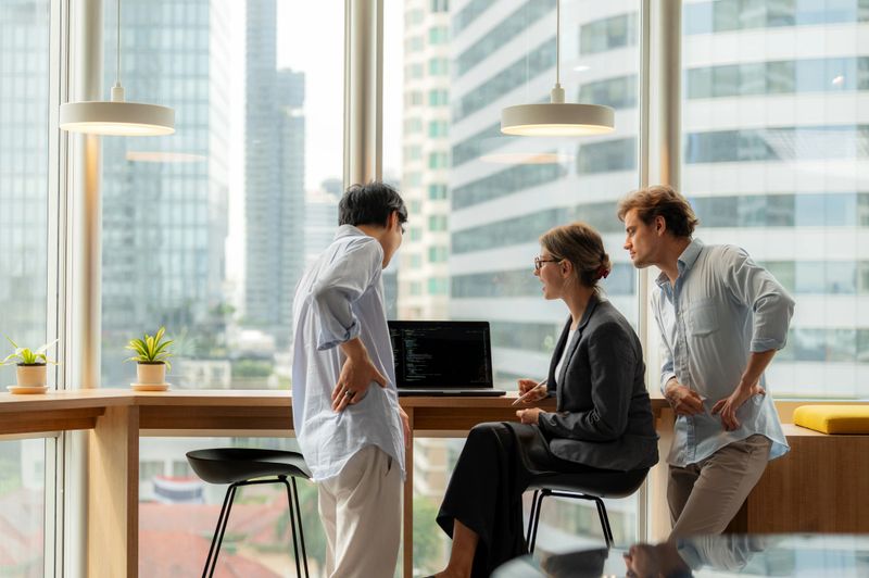 A group of business people from different backgrounds converse in a bright office setting by floor-to-ceiling windows. The dynamic scene emphasizes innovation, strategy, communication, and professional teamwork in a high-rise business district.