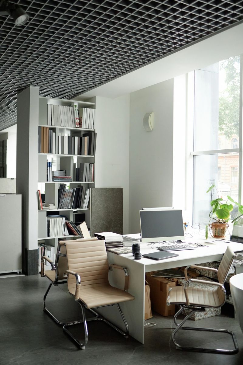 Modern office workspace featuring empty chairs surrounding desk with computer monitor, bookshelves filled with documents in background, large window letting in natural light, no people visible