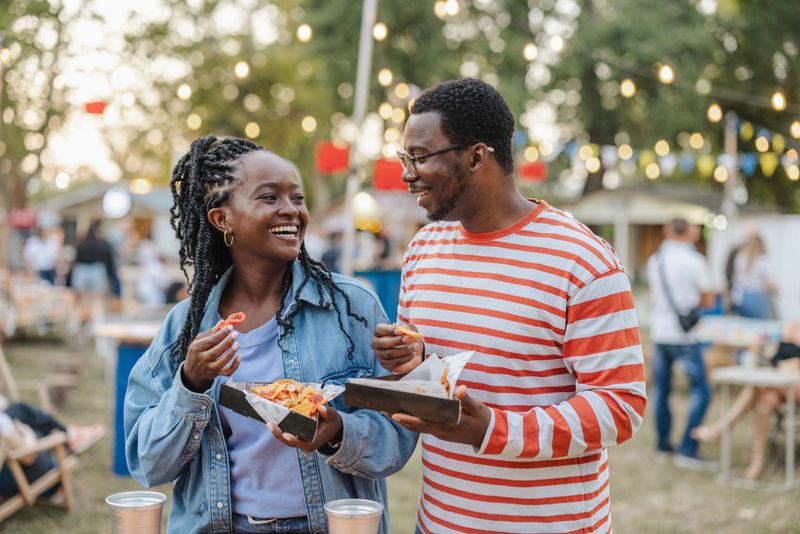Two cheerful individuals share a moment while enjoying delicious food at an open-air festival. The backdrop includes festive lights, greenery, and other attendees, conveying a lively and relaxed atmosphere.