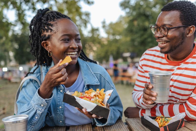 Two friends are spending time together outdoors, enjoying snacks and drinks at a picnic table.