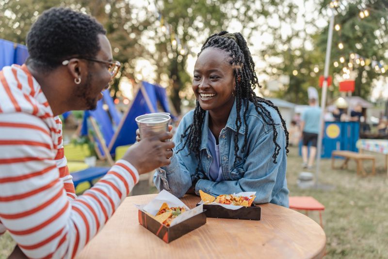 Two friends share laughter and casual dining in an outdoor festival area surrounded by vibrant booths.