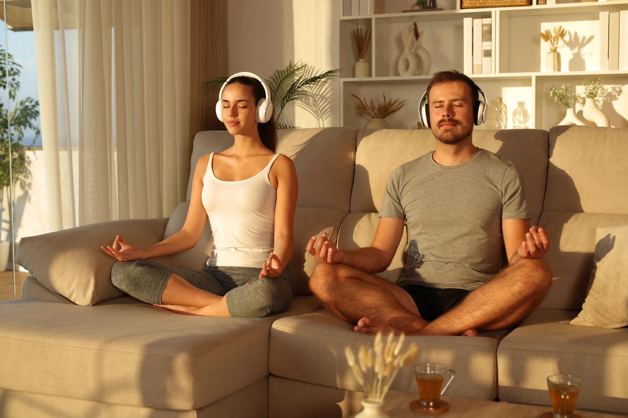 Couple of amateur yogis doing yoga at home listening online guide from headphones sitting on a sofa at sunset