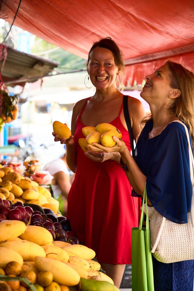 Two women cheerfully selecting ripe mangoes at a colorful and lively market under a bright canopy.