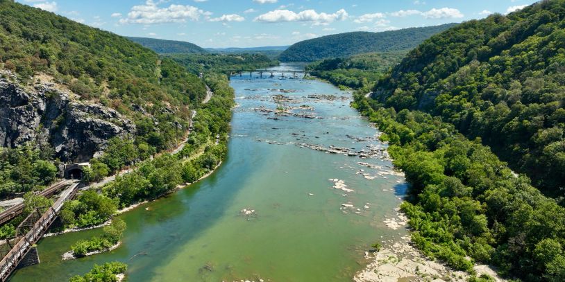 A wide river flanked by green hills and rocky cliffs under a partly cloudy sky.