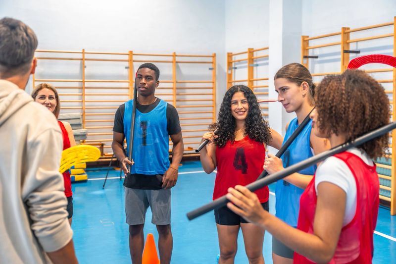 Multi-ethnic group of young adult athletes gathered inside a gym, attentively listening to their coach before an intense floorball training session, focused on skill improvement and teamwork