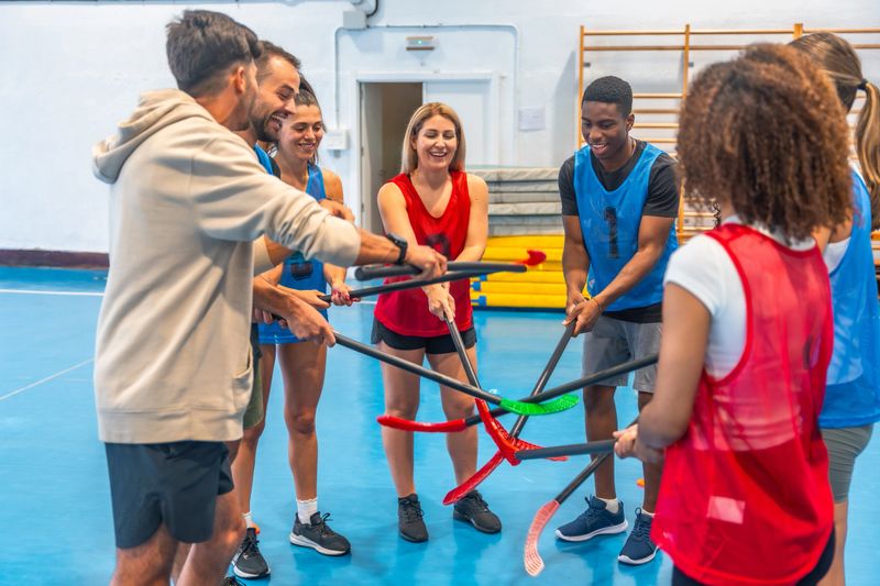 Group of young athletes forming a circle in a gym, holding their floorball sticks together, smiling and looking at each other, getting ready to play
