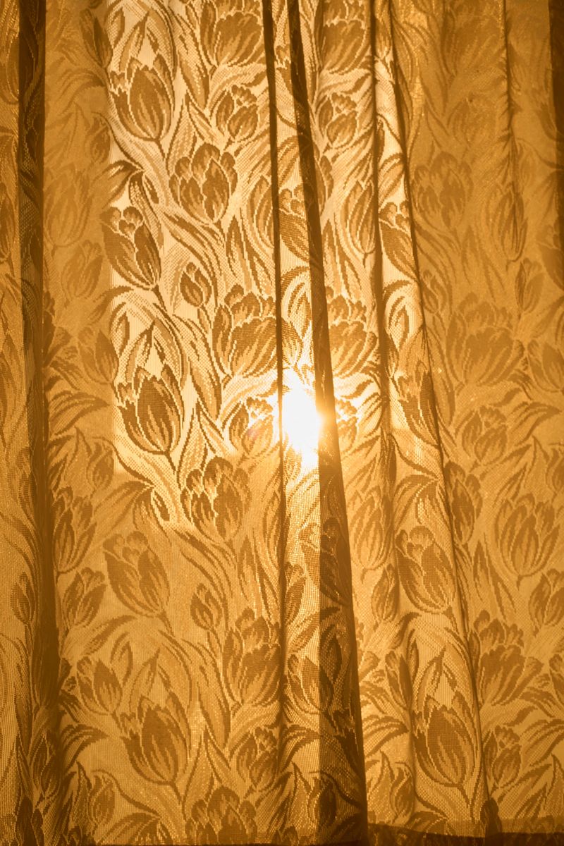 Horizontal interior scene of vintage floral curtains with a decorative tassel trim, photographed backlit as warm sunlight glows through the patterned fabric. The folds and pleats create texture and a nostalgic, cozy atmosphere suitable for themes of home, privacy, hospitality, and morning calm. Soft golden tones provide ample copy space for titles or branding. Works for editorial features, lifestyle and décor content, brochures, ads, and website hero images. No people and no visible trademarks; full frame.