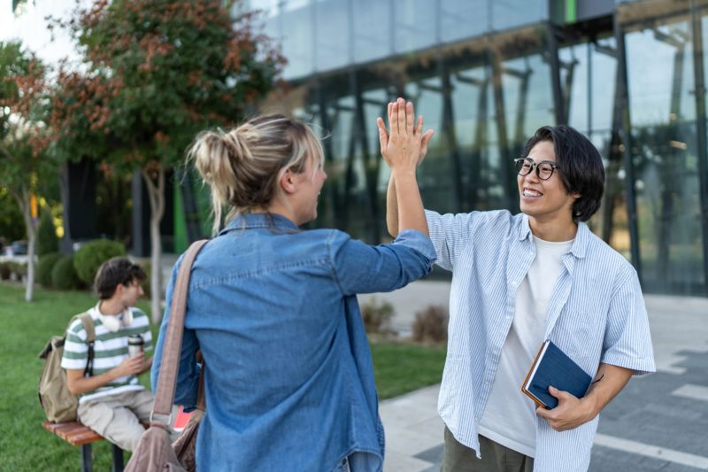 Two happy, multi-ethnic students sharing a high five outdoors on a university campus.