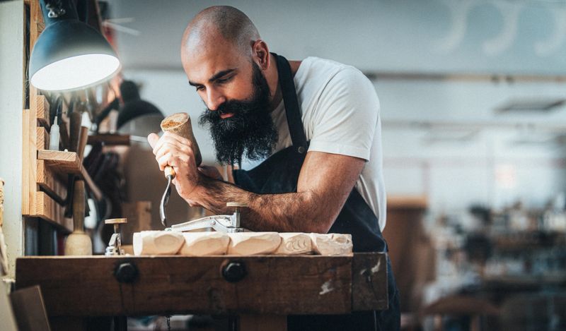 Craftsman deeply focused on woodworking in workshop. The man has a bald head and a thick, dark beard, and he is wearing a white t-shirt under a blue apron with leather accents. He is carefully carving a piece of wood using a chisel and a wooden mallet, showing precision and concentration.The workspace around him is well-organized, with various woodworking tools.