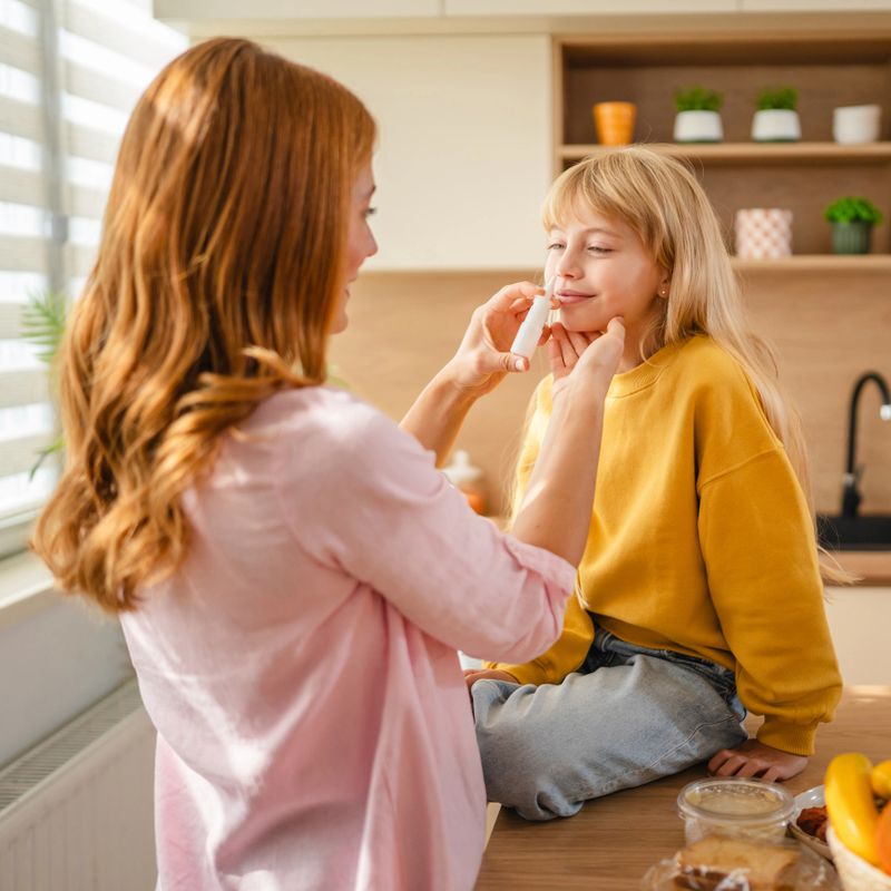 Mother gently applying nasal spray to her daughter's nose in their cozy kitchen, offering comfort and relief during a time of illness and allergy struggles