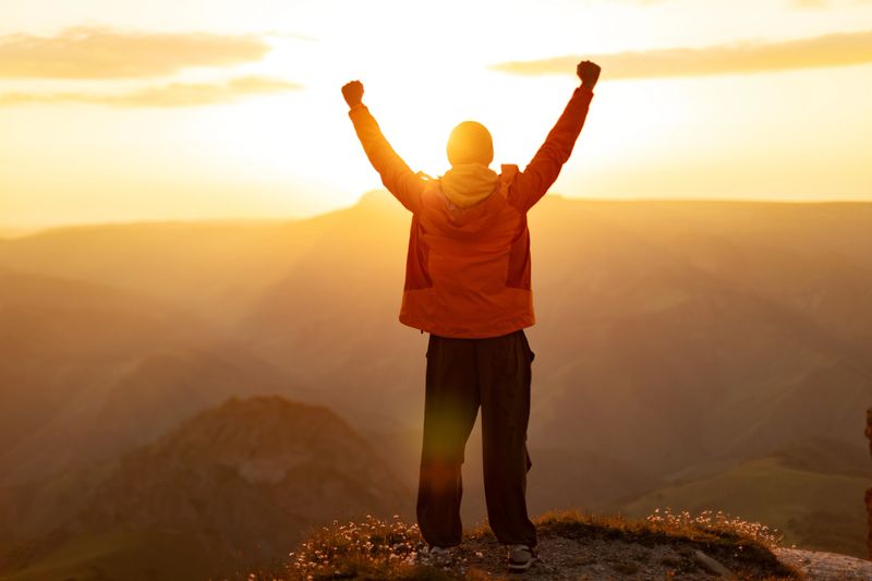 Mid adult man stands on mountain summit with arms raised, looking at sunset and celebrates success