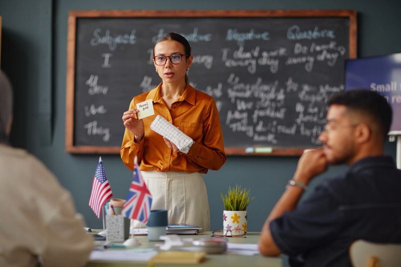 Caucasian young adult woman teaching language lesson holding flashcard and notebook, standing in front of chalkboard while multiethnic young adults sitting at table listening attentively