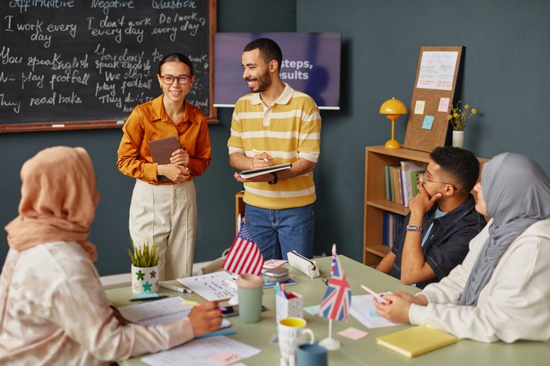 Diverse group of young adults participating in language school lesson, two instructors standing and smiling while students sitting at table with notebooks and flags