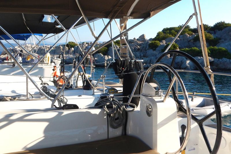 Close view of a sailing yacht cockpit with wheel, ropes and bimini frames, moored near rocky islets on blue water under summer light. Clean marine scene for travel, leisure, charter and lifestyle use