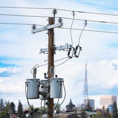Close-up of electrical transformers on a utility pole with city skyline in the background.