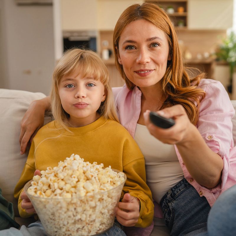 Happy mother and daughter sitting on a cozy sofa, enjoying a movie night with popcorn and laughter, creating a heartwarming scene of family bonding