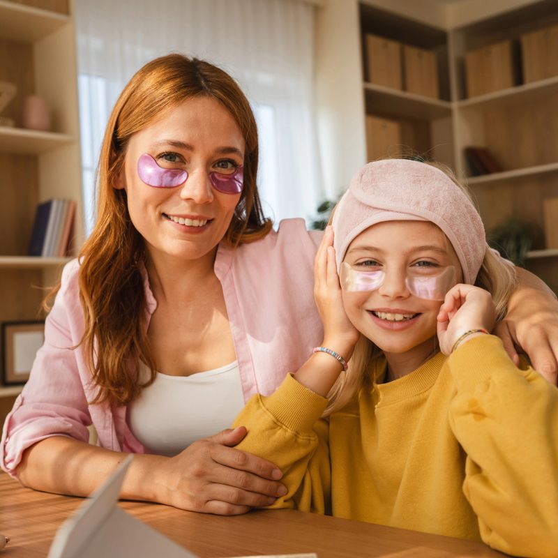Mother and daughter are wearing hydro gel patches under their eyes while sitting at a table in their living room, enjoying a moment of pampering and bonding together
