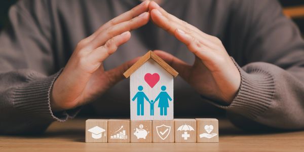 Hands protecting a family home model with insurance icons.