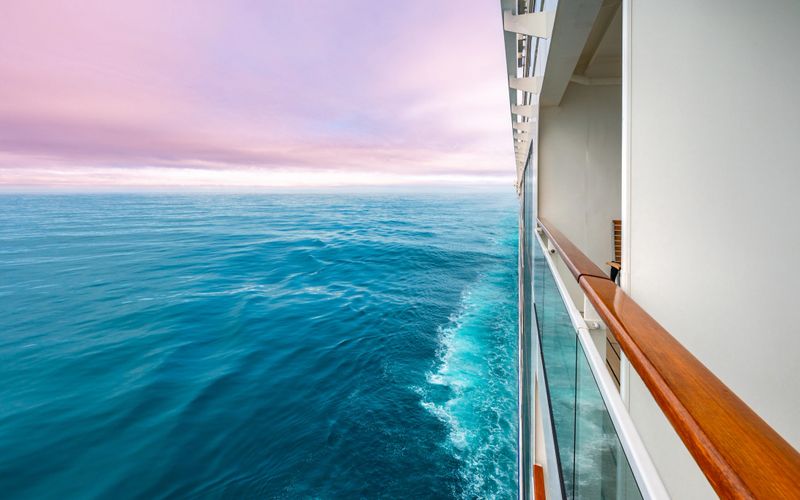 Colorful travel photo of a cruise ship sailing on the ocean with a view of the horizon and a pink-toned sunset sky. Perfect for themes of vacation, adventure, and maritime travel.