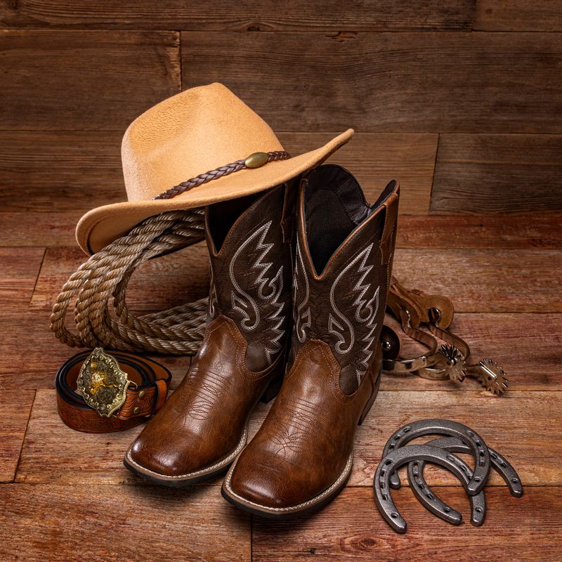 Still life studio shot featuring a pair of cowboy boots, a white cowboy hat, coiled rope, and a large belt buckle arranged together on a retro wooden background. Western Americana theme with rustic vintage styling and country lifestyle details.