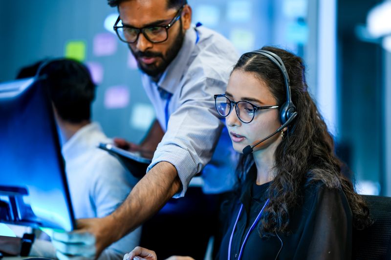 A team leader trains a new customer service agent in a modern call center. The diverse team works together to provide professional and helpful technical support to clients.