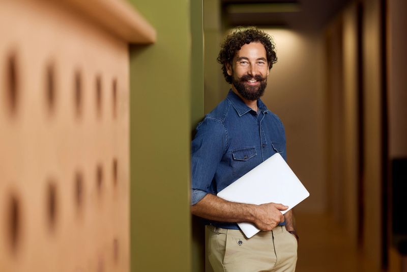 A cheerful man holding a laptop smiles while standing against a wall indoors.