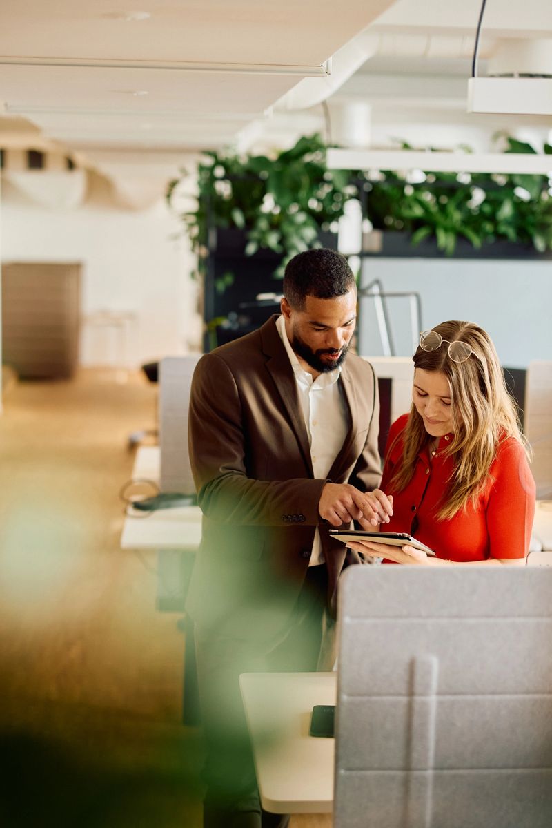 Business man in a suit and woman in a red blouse standing together, using a tablet together, surrounded by workstations and greenery in a contemporary office.