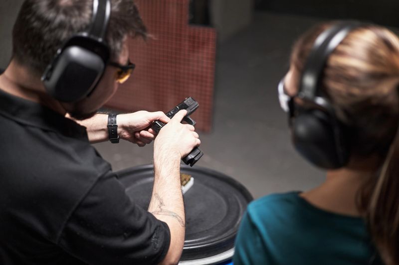 Caucasian middle aged man instructing Caucasian young woman on handgun handling at indoor shooting range, both wearing protective ear muffs and safety glasses, focusing on firearm safety
