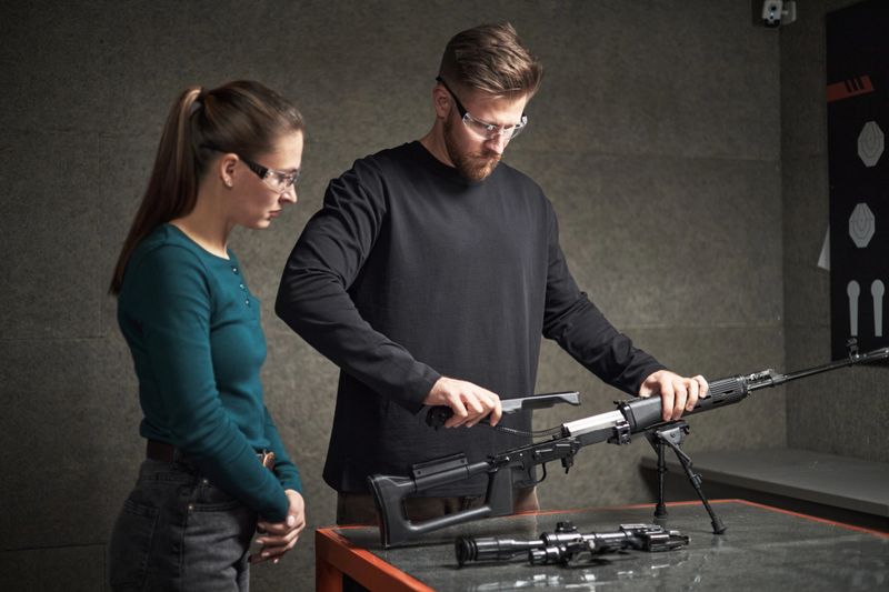 Caucasian young adult man demonstrating rifle assembly to Caucasian young adult woman in indoor shooting range, both wearing protective eyewear and standing at table with firearm parts