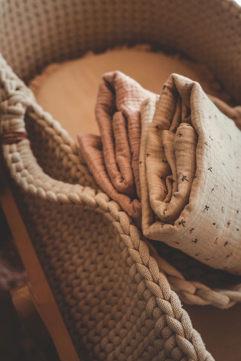 Cozy close-up of soft knitted baby blanket and muslin fabric with floral pattern, placed in woven basket on wooden floor, warm natural lighting, nursery decor.