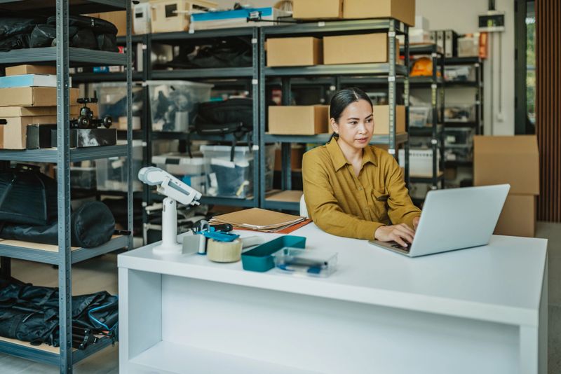 In a well-organized warehouse, a young Asian woman works diligently on her laptop, managing inventory and processing orders to streamline operations for her retail business.