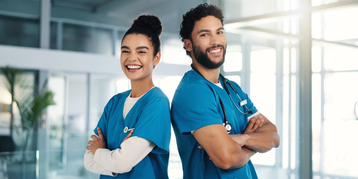 Two smiling healthcare workers in blue scrubs standing back to back.