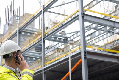 Construction worker in a yellow jacket and white helmet talks on phone at site.