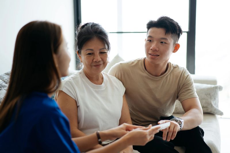 A healthcare professional speaks with an older woman and a younger man during a home visit. The provider holds a device while discussing care instructions. The image highlights home support, health guidance, and family involvement in medical decision-making.