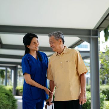A caregiver in blue scrubs assisting an elderly man outdoors.