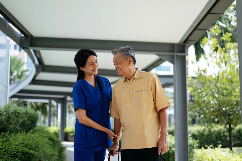 A healthcare professional supports an elderly man as he walks with a cane along a covered walkway. The nurse smiles warmly, demonstrating caregiving and encouragement during outdoor rehabilitation