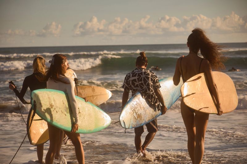 Surfers head towards the ocean carrying their boards, ready to catch waves on a sunny day. The scene captures the joy, camaraderie, and energy associated with surfing and coastal activities.