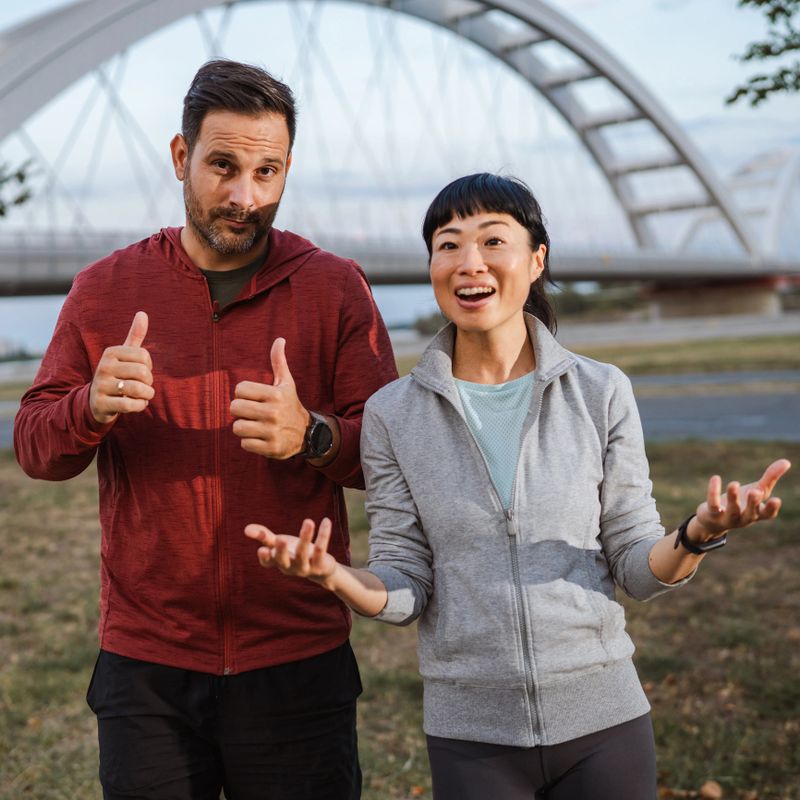 Man and woman wearing sporty attire are gesturing with their hands, engaging in a lively discussion while enjoying an outdoor workout near a scenic bridge during sunset