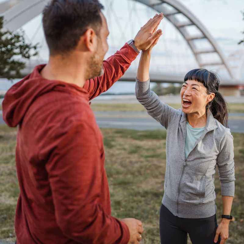 Smiling couple of joggers is giving a high five after running near a bridge in an urban environment, celebrating their achievement and healthy lifestyle