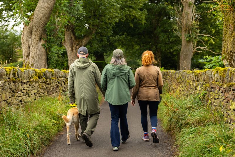 A visually impaired man in his 60s  walking with his wife, daughter and his guide dog through a scenic rural path on an overcast day.