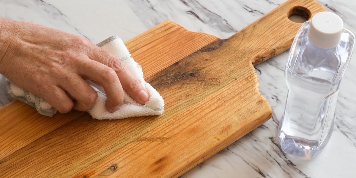 Hand wiping wooden cutting board with cloth beside water bottle.